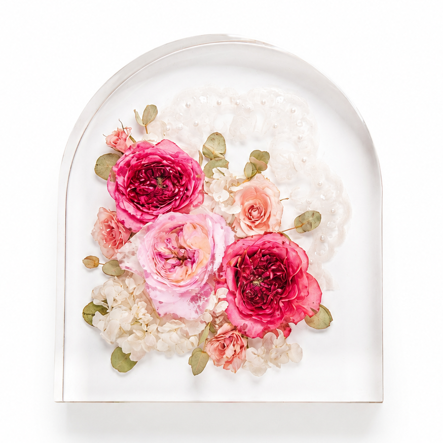 An arched clear acrylic wall plaque featuring a pressed floral arrangement of pink and peach roses, cream hydrangeas, and green leaves, set against a white background.