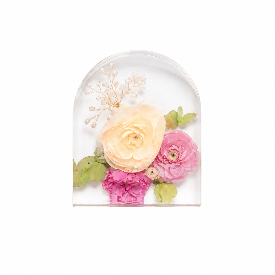 A clear arched resin dome containing a pressed floral arrangement with a large pale peach rose, pink ranunculus, green leaves, and delicate beige dried stems, centered on a white background.